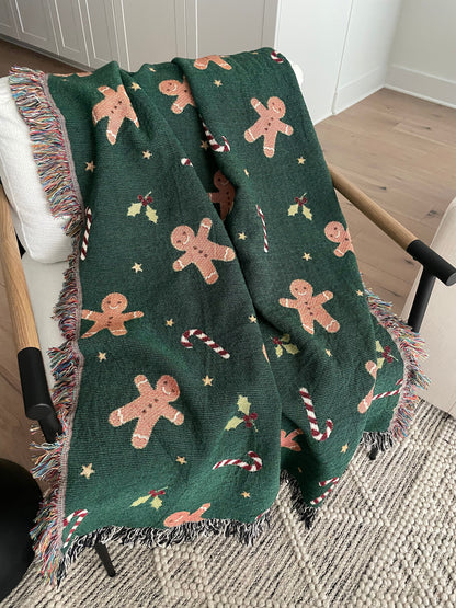 Dark green Christmas woven blanket featuring gingerbread men, candy canes, holly leaves, and multicolored plus black-and-white fringe, draped over a white armchair.