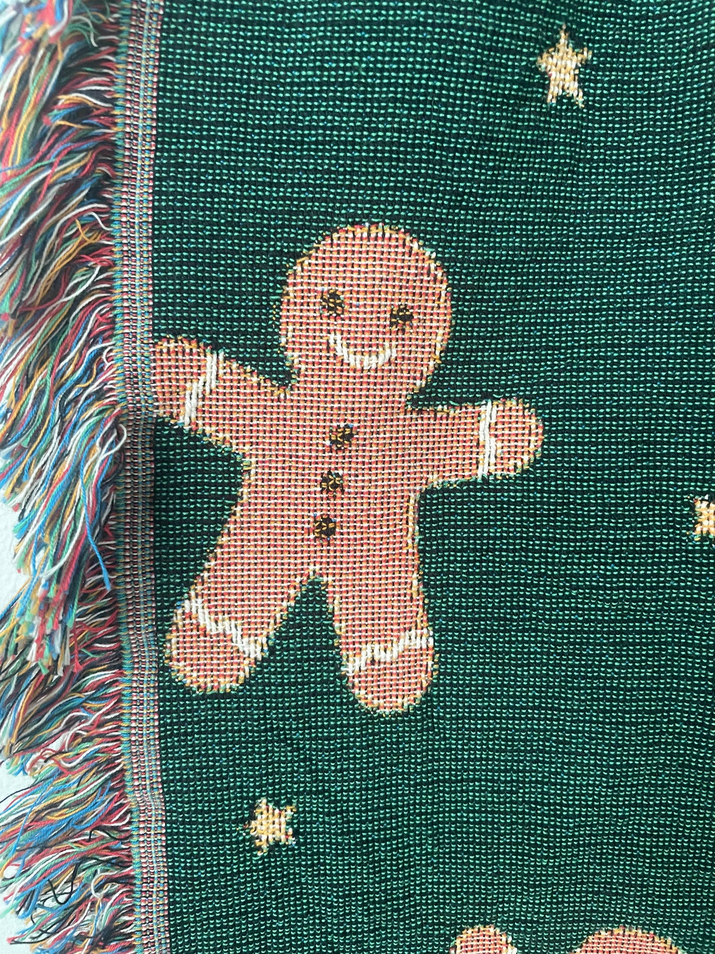 Close-up of a dark green Christmas woven blanket showing a gingerbread man motif, small woven stars, and multicolored fringe along the edge.