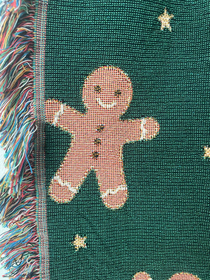 Close-up of a dark green Christmas woven blanket showing a gingerbread man motif, small woven stars, and multicolored fringe along the edge.
