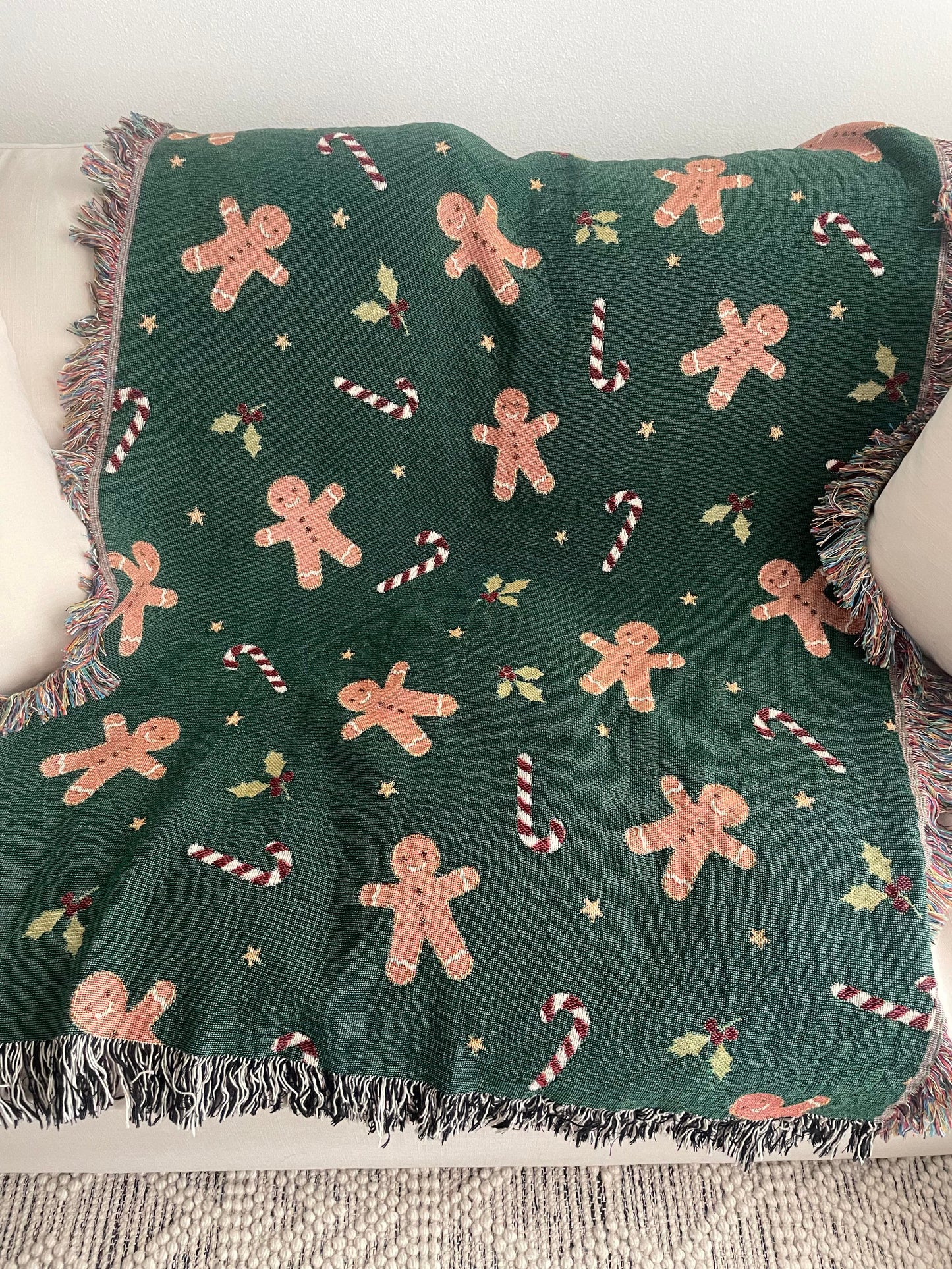 Dark green Christmas woven blanket featuring gingerbread men, candy canes, holly leaves, and multicolored plus black-and-white fringe, spread across a cream-colored sofa.