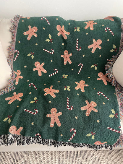 Dark green Christmas woven blanket featuring gingerbread men, candy canes, holly leaves, and multicolored plus black-and-white fringe, spread across a cream-colored sofa.