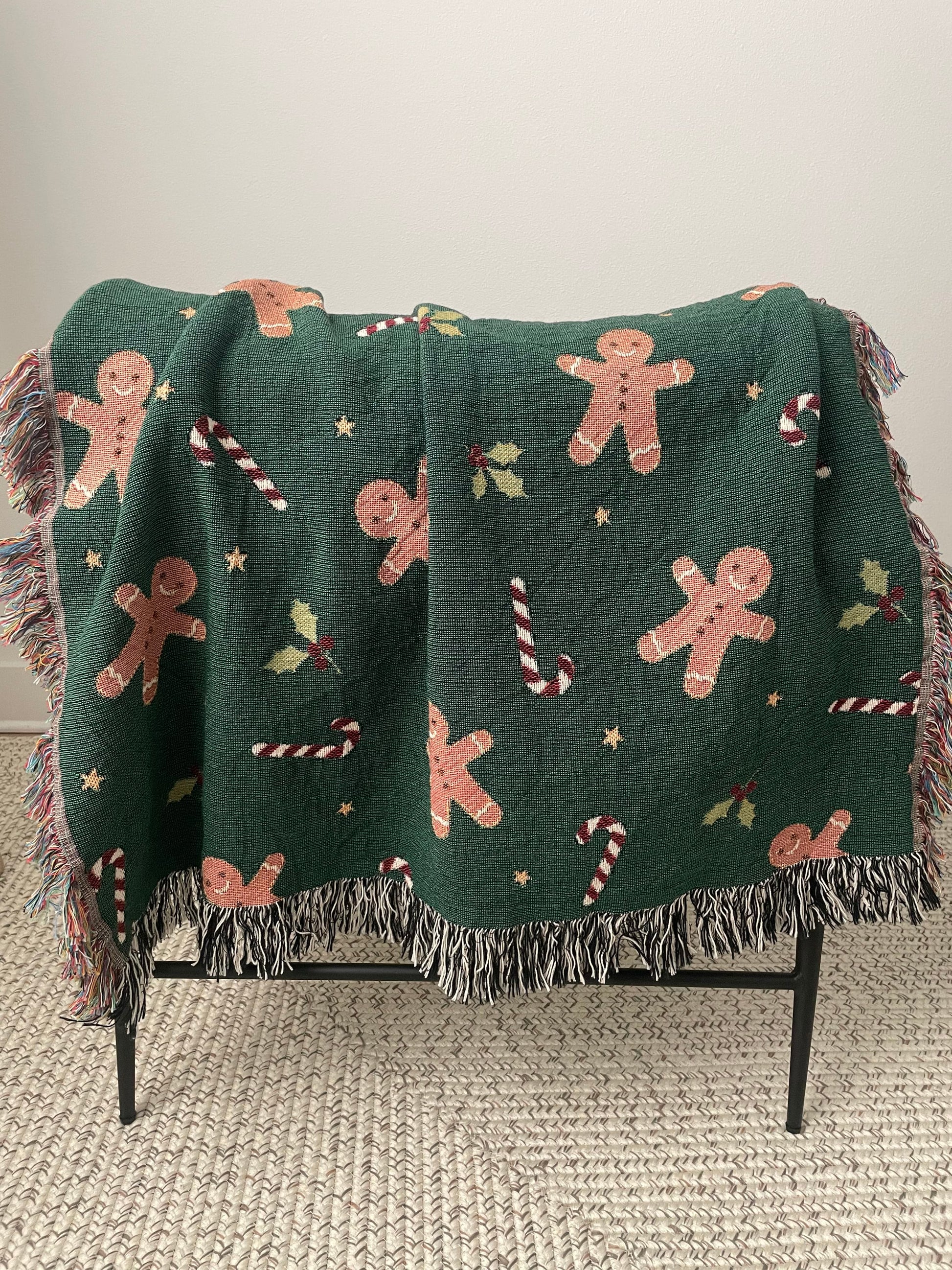 Dark green Christmas woven blanket featuring gingerbread men, candy canes, holly leaves, and multicolored plus black-and-white fringe, draped over the back of a modern armchair.