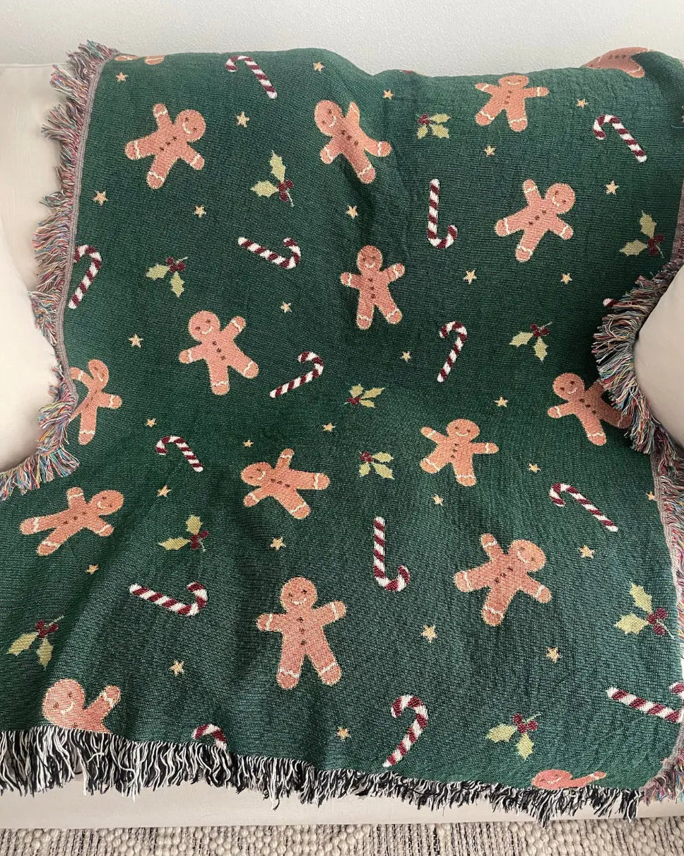 A dark green woven throw blanket with a festive holiday pattern of gingerbread men, candy canes, holly, and stars, draped over a white sofa.