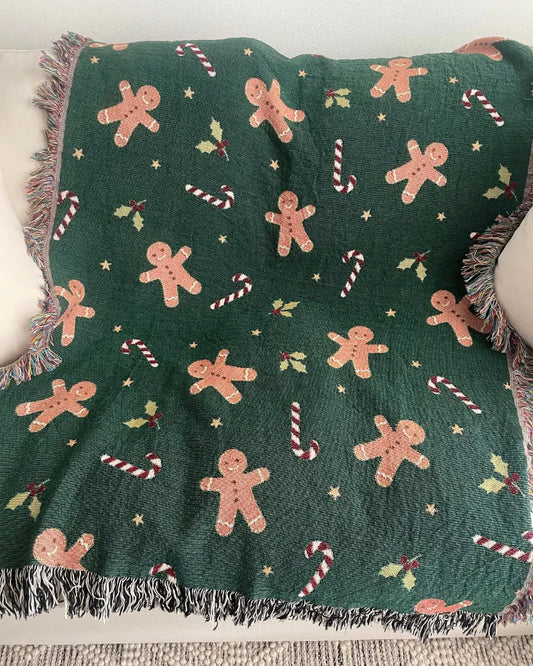 A dark green woven throw blanket with a festive holiday pattern of gingerbread men, candy canes, holly, and stars, draped over a white sofa.