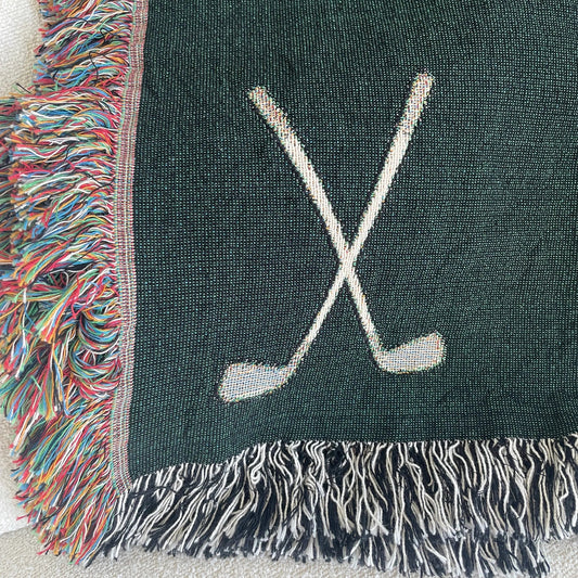 Close-up of forest green golf clubs woven throw blanket showing crossed golf club motif, golf ball accent, and fringe edge detail