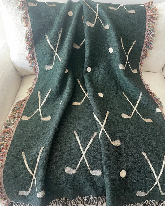 Golf woven throw blanket in forest green with an all-over pattern of crossed golf clubs and golf balls, finished with black-and-white and multicolored fringe and draped over a white sofa.