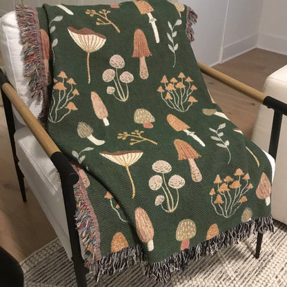 A green woven throw blanket with mushroom and botanical motifs, featuring fringed edges, draped over a chair.

