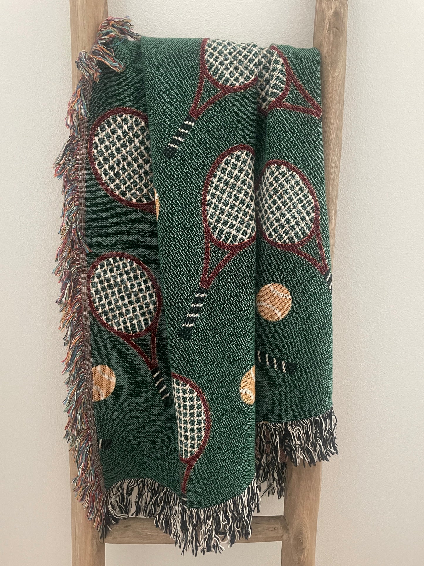 Green tennis racket woven throw blanket with fringe displayed on a wooden ladder.