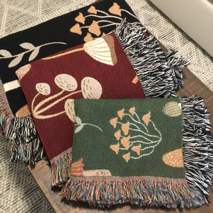 Mushroom-themed woven throw blankets in black, red, and green, featuring multicolor fringe on one end and black-and-white fringe on the other, neatly folded and display on a wooden bench.

