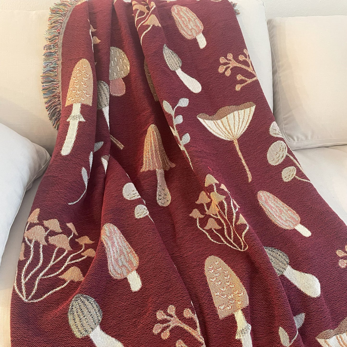 Red woven throw blanket featuring detailed mushroom and foliage pattern, draped over a cream couch — perfect for cottagecore or fall decor.