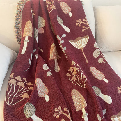 Red woven throw blanket featuring detailed mushroom and foliage pattern, draped over a cream couch — perfect for cottagecore or fall decor.