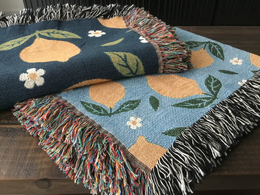 Close-up of lemon and daisy patterned woven throw blankets with fringe edges, in navy and light blue, folded and layered on a rustic wooden table.