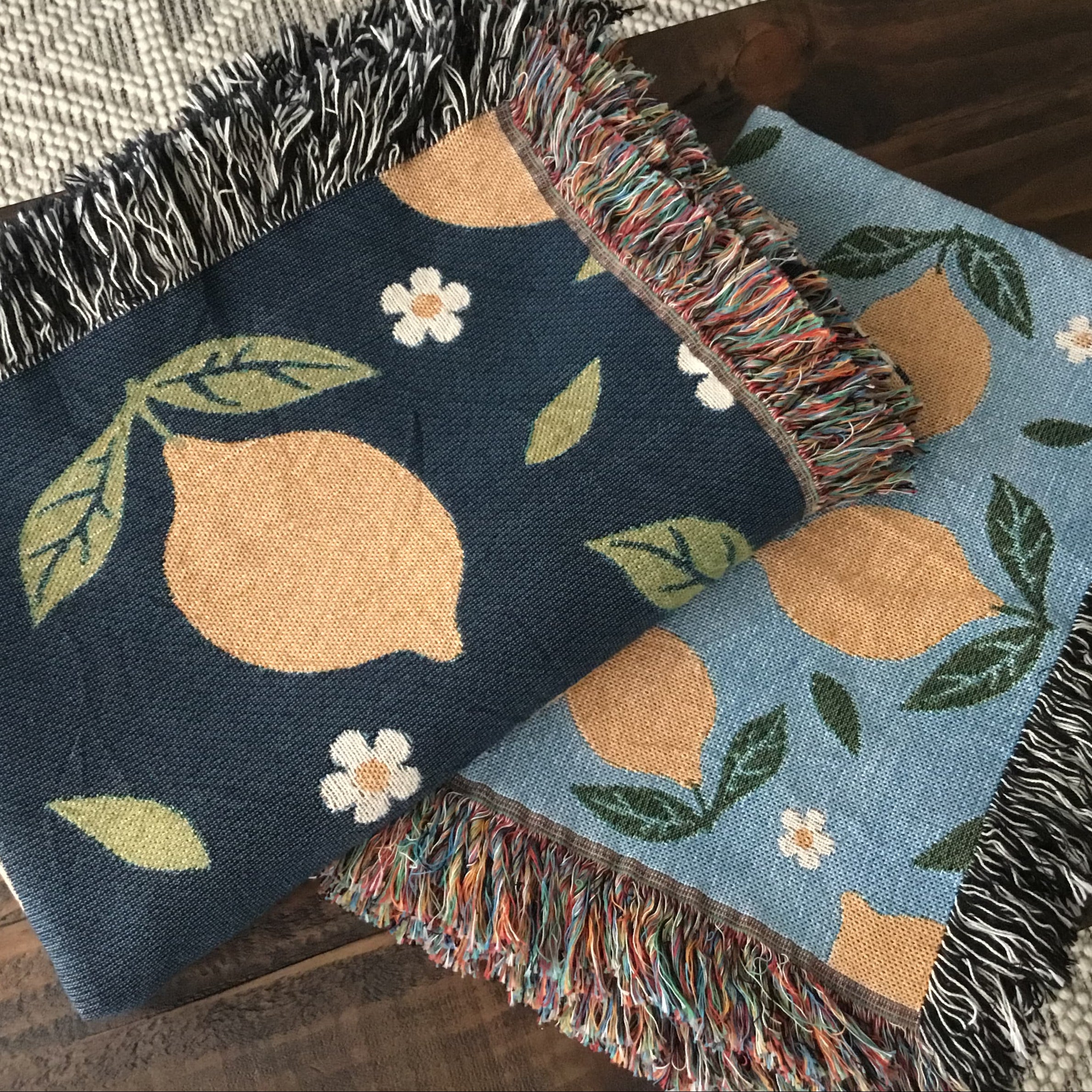 Two lemon-themed woven throw blankets with daisy patterns—one in navy and one in light blue—featuring fringe edges, folded on a wooden surface.