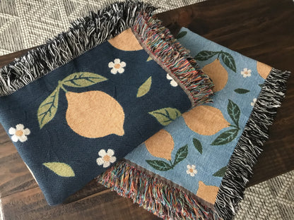 Two lemon-themed woven throw blankets with daisy patterns—one in navy and one in light blue—featuring fringe edges, folded on a wooden surface.