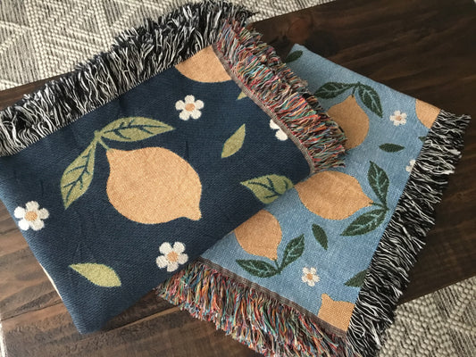 Two lemon-themed woven throw blankets with daisy patterns—one in navy and one in light blue—featuring fringe edges, folded on a wooden surface.