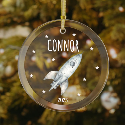 Personalized rocket Christmas glass ornament with custom name and year, featuring a silver rocket ship and stars. A unique holiday gift for space-loving kids and adults.
