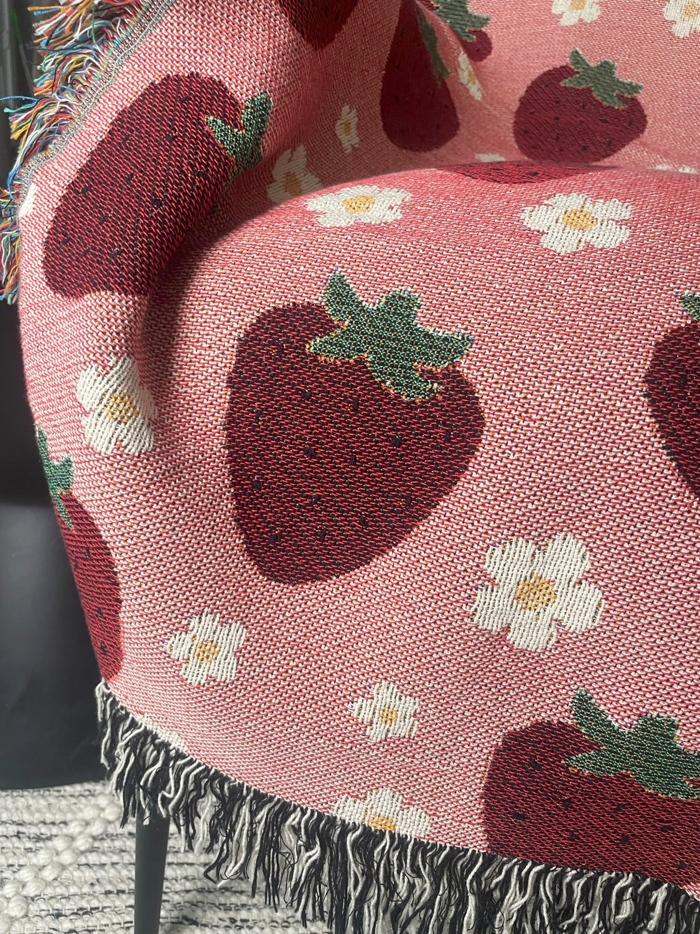 Close-up of pink woven strawberry and daisy throw blanket with multicolored and black-and-white fringe, draped over a chair for cozy cottagecore home decor.