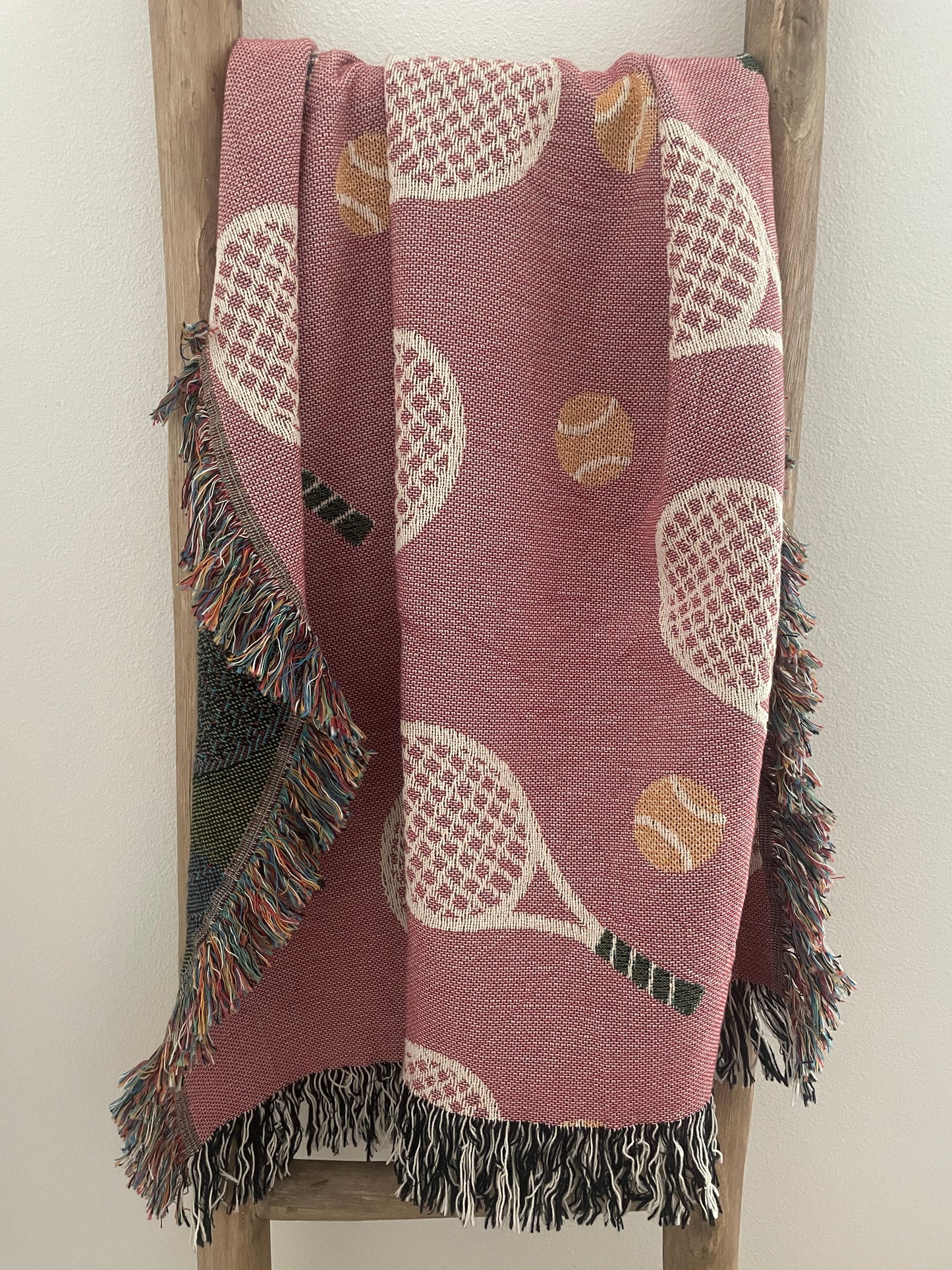 Pink tennis racket woven throw blanket with fringe displayed on a wooden ladder.