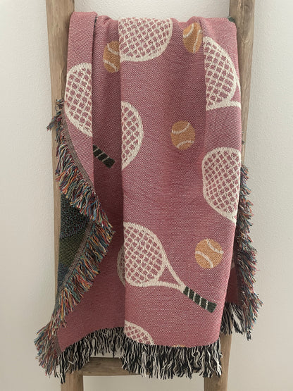 Pink tennis racket woven throw blanket with fringe displayed on a wooden ladder.
