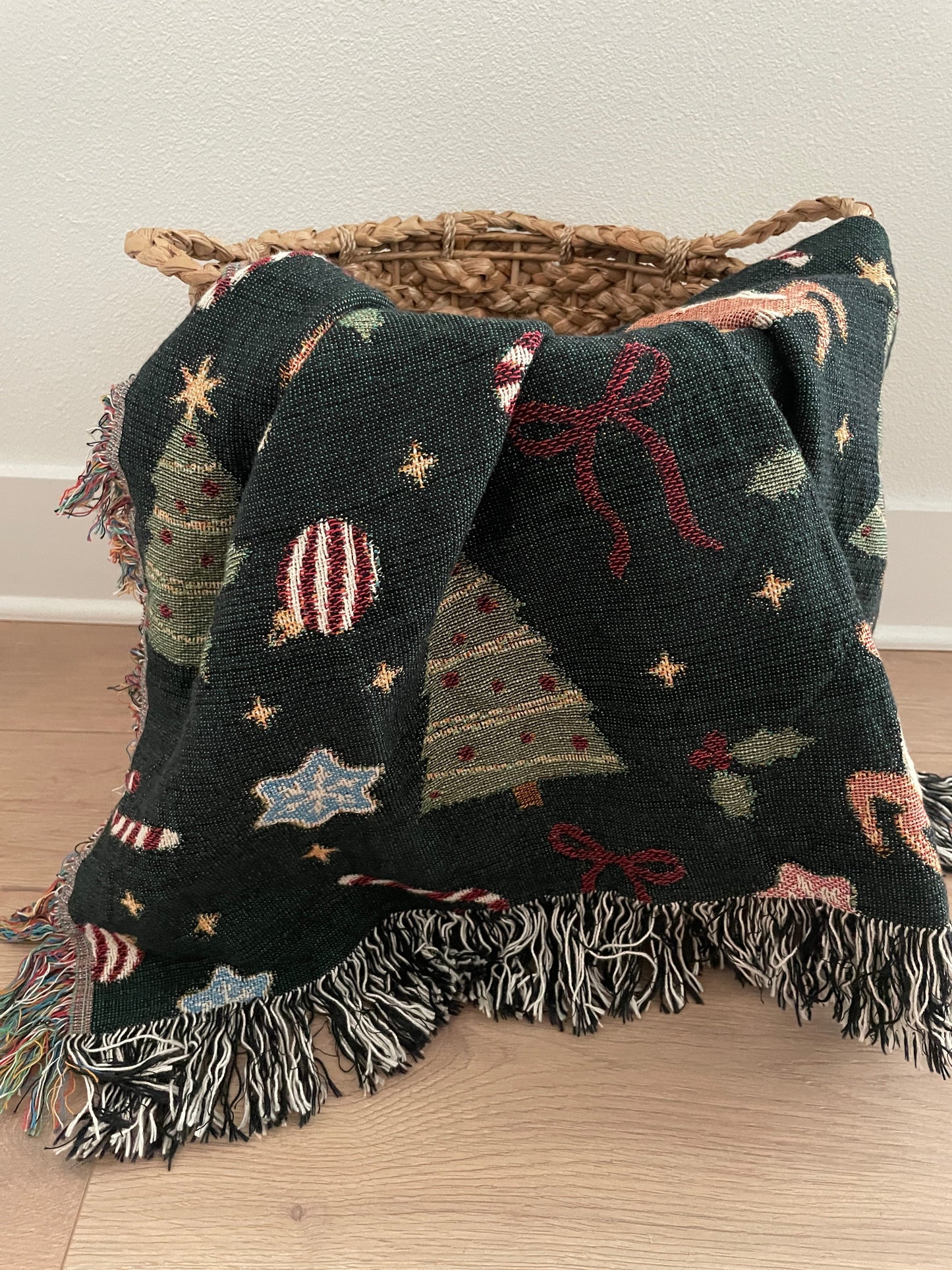 Dark green woven Christmas throw blanket with holiday motifs including reindeer, Christmas trees, bows, stars, holly, and ornaments, styled draped over a wicker basket with fringe edges.