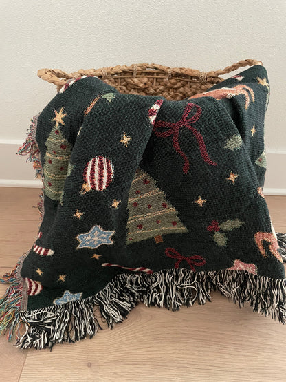Dark green woven Christmas throw blanket with holiday motifs including reindeer, Christmas trees, bows, stars, holly, and ornaments, styled draped over a wicker basket with fringe edges.