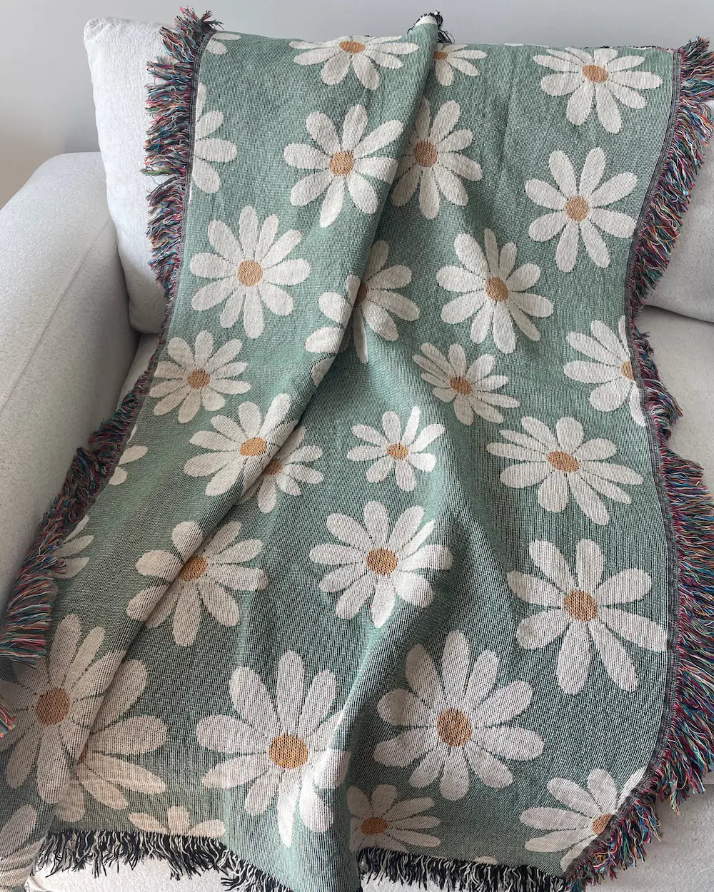 Sage green daisy woven throw blanket with fringed edges, draped over a white sofa.