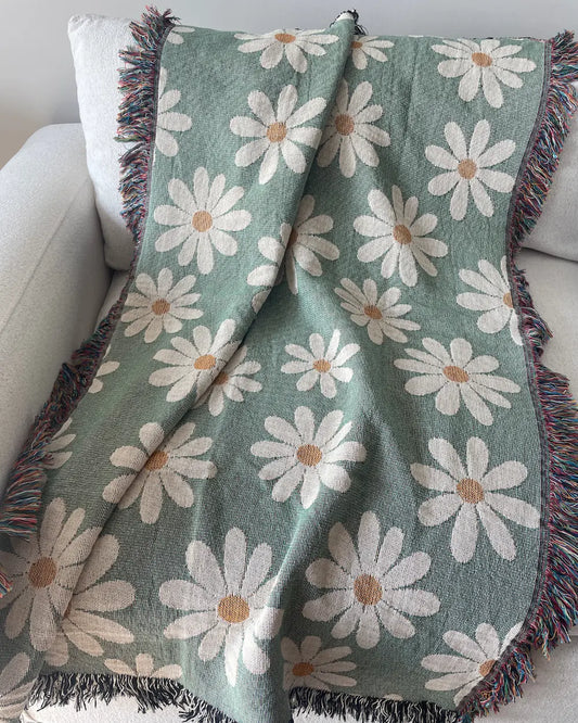 Sage green daisy woven throw blanket with fringed edges, draped over a white sofa.