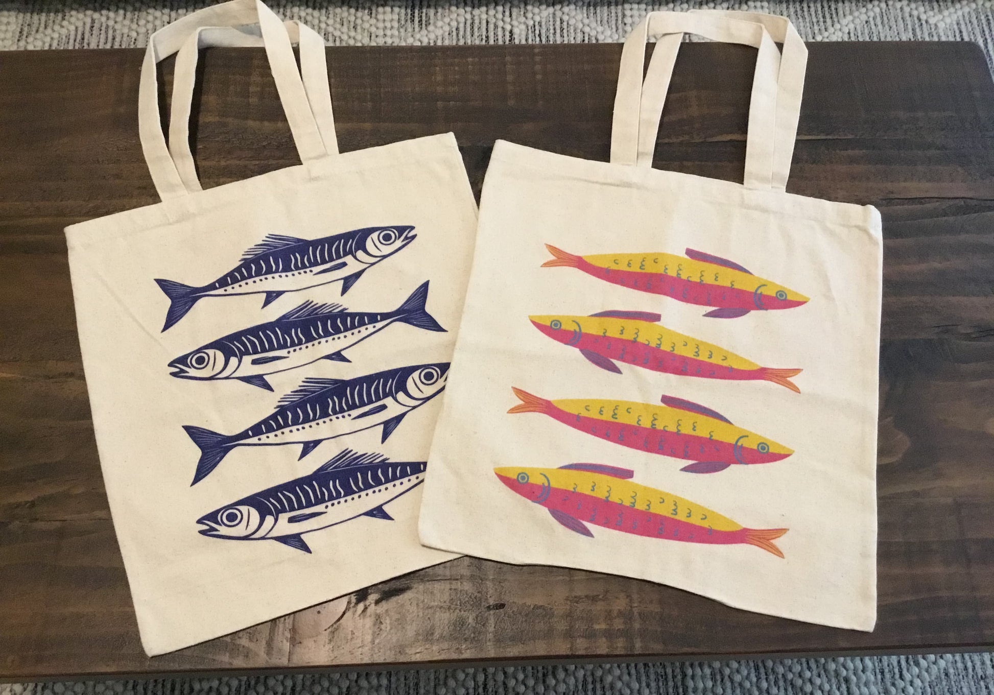 Two sardine-print cotton canvas tote bags on table – one with navy blue fish, one with colorful yellow and pink fish, perfect for coastal style or gifts.