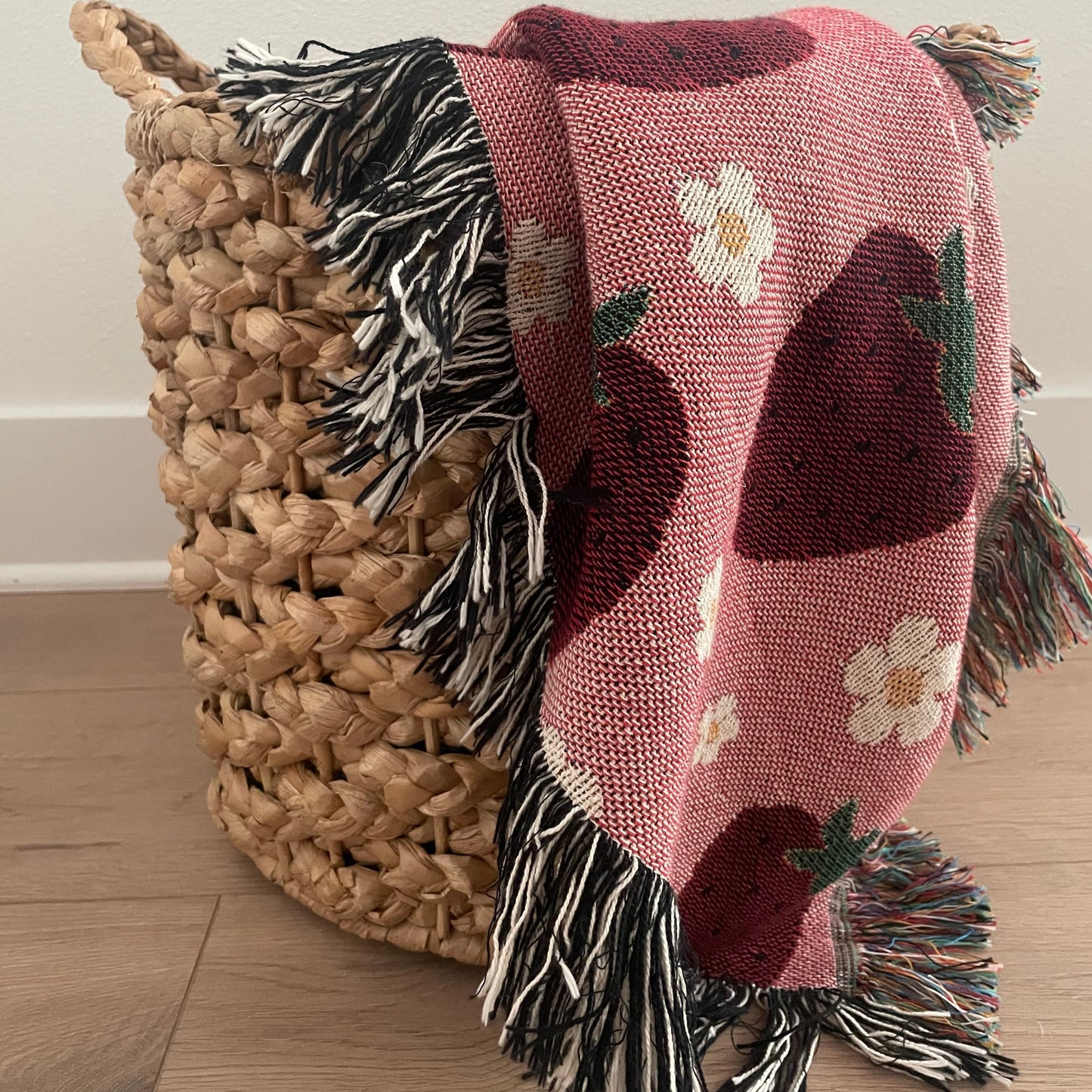 Pink woven strawberry and daisy throw blanket draped over a natural wicker basket on a light wood floor, featuring multicolored and black-and-white fringe