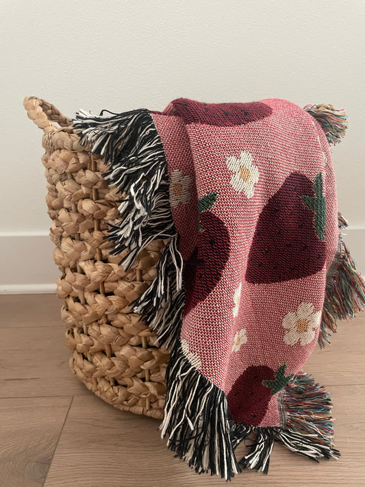 Pink woven strawberry and daisy throw blanket draped over a natural wicker basket on a light wood floor, featuring multicolored and black-and-white fringe