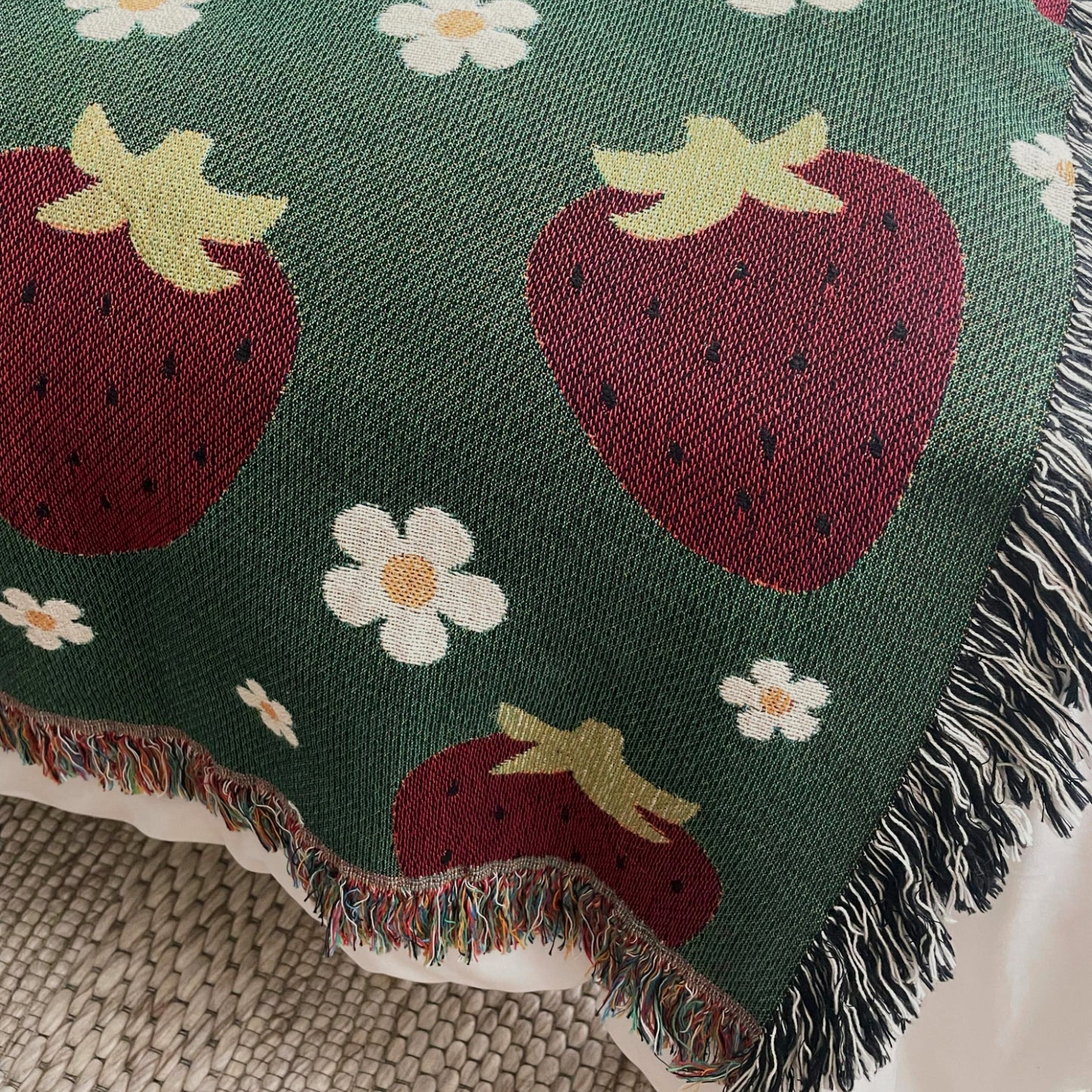 Green woven throw blanket with red strawberries and white daisies, featuring multicolored and black-and-white fringe, styled on a bed for cottagecore or farmhouse decor.