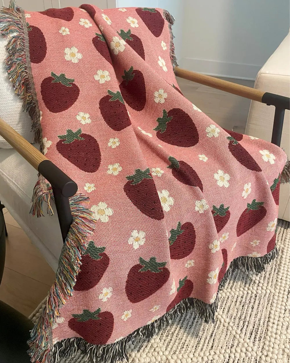 A pink woven throw blanket with a red strawberry and white daisy flower pattern, draped over a wooden armchair in a living room setting.