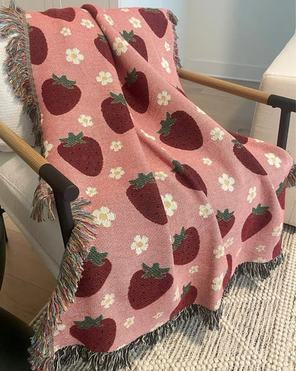 A pink woven throw blanket with a red strawberry and white daisy flower pattern, draped over a wooden armchair in a living room setting.