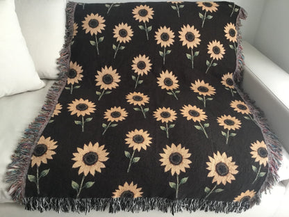 Dark brown woven sunflower throw blanket laid flat over a white couch, showcasing a repeating yellow sunflower pattern and multicolored and black and white fringe edges.









