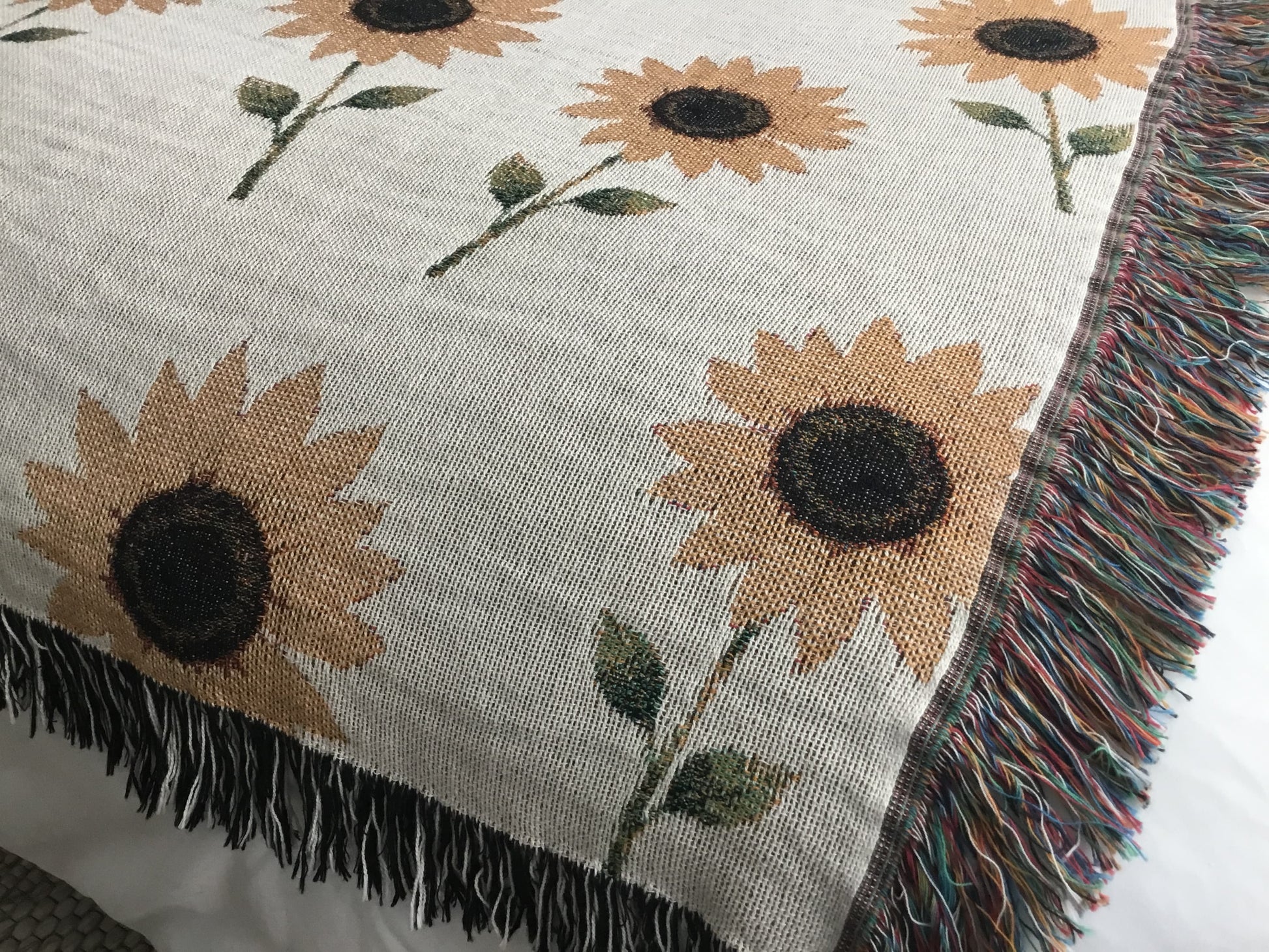 Close-up of a beige woven sunflower throw blanket featuring a repeating pattern of sunflower plants with dark brown centers, green leaves, and stems, finished with multicolored fringe on the sides and black-and-white fringe along the top and bottom edges.