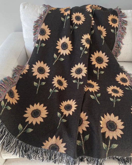 Sunflower woven throw blanket with golden yellow flowers and green leaves on a dark brown background, finished with black-and-white and multicolored fringe and draped over a white sofa.
