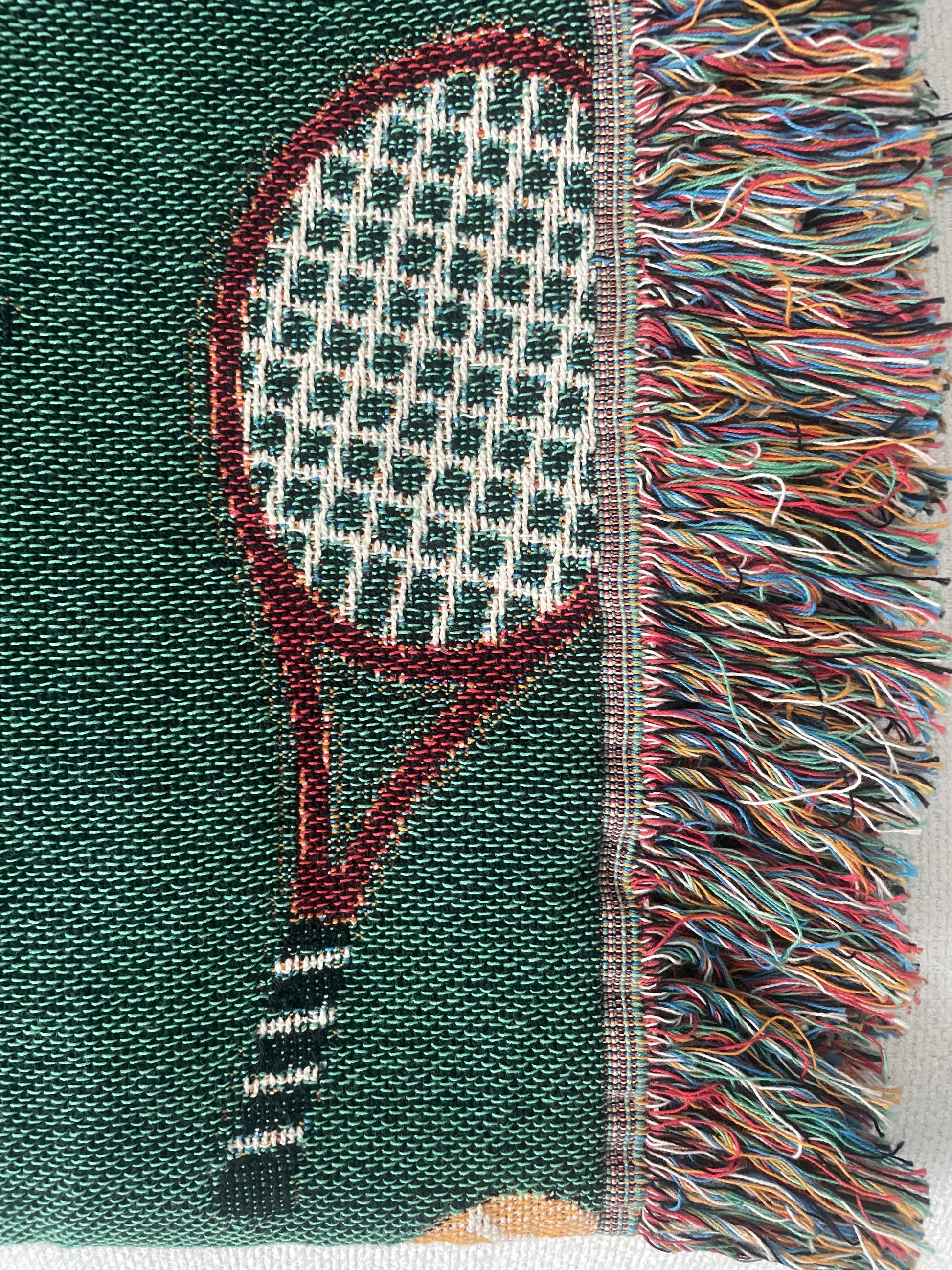 Close-up of a green woven tennis racket throw blanket showing detailed stitching and multicolor fringe.