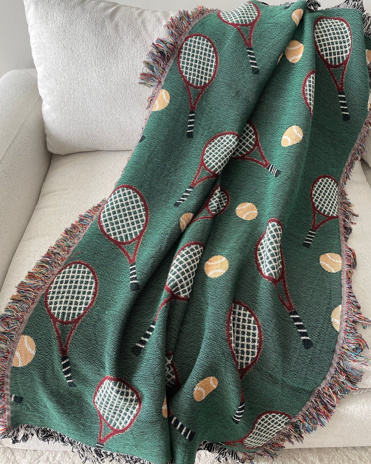 Green woven throw blanket with a repeating tennis racket and tennis ball
pattern, draped over a light beige sofa with fringe edges.