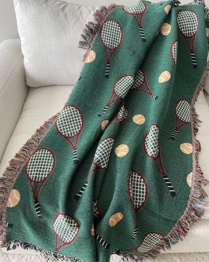 Green woven throw blanket with a repeating tennis racket and tennis ball
pattern, draped over a light beige sofa with fringe edges.