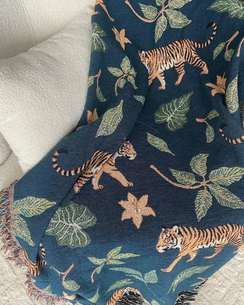Dark teal-blue woven throw blanket with walking tigers, green tropical leaves, orange flowers, and fringed edges draped over a white sofa.