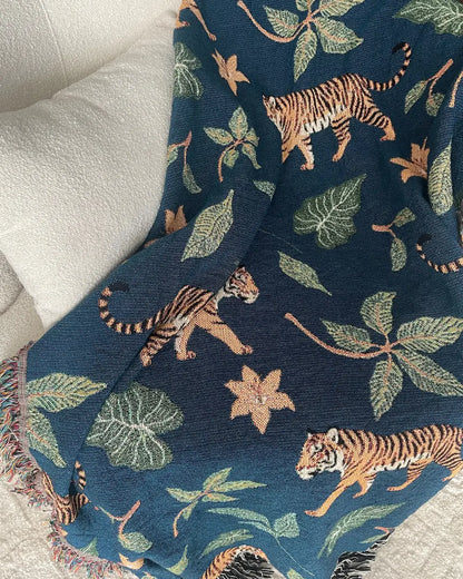 Dark teal-blue woven throw blanket with walking tigers, green tropical leaves, orange flowers, and fringed edges draped over a white sofa.