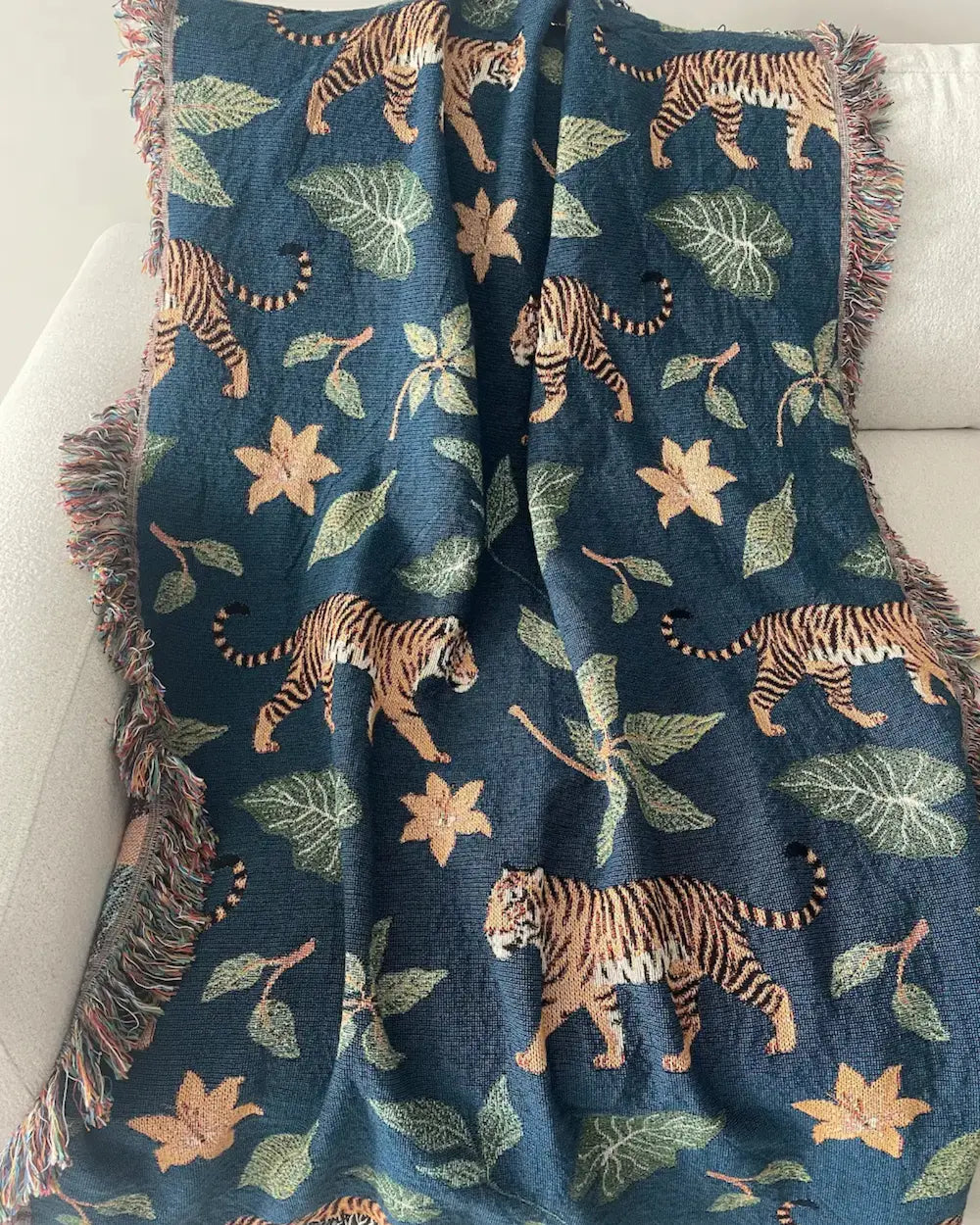 Woven throw blanket draped over a white sofa, featuring a dark teal-blue background with walking tigers, green tropical leaves, orange flowers, and fringed edges.