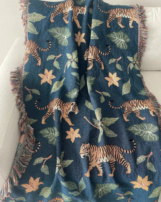 Woven throw blanket draped over a white sofa, featuring a dark teal-blue background with walking tigers, green tropical leaves, orange flowers, and fringed edges.