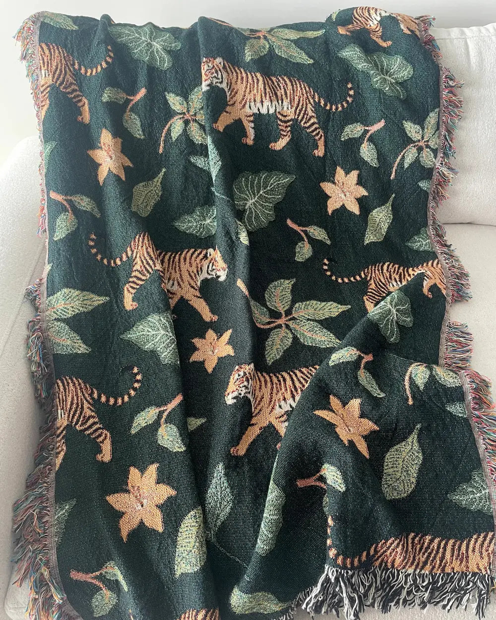 Dark green woven throw blanket with walking tigers, tropical leaves, orange flowers, and fringed edges draped over a white sofa.