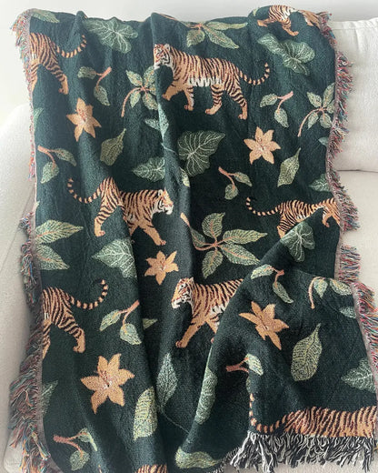Dark green woven throw blanket with walking tigers, tropical leaves, orange flowers, and fringed edges draped over a white sofa.