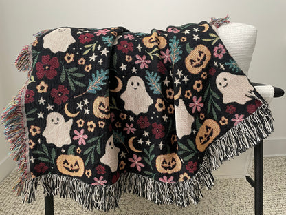 Halloween-themed woven throw blanket draped over a white chair, featuring a playful pattern of smiling ghosts, jack-o’-lanterns, crescent moons, stars, and florals. Finished with multicolor and black-and-white fringe edges.